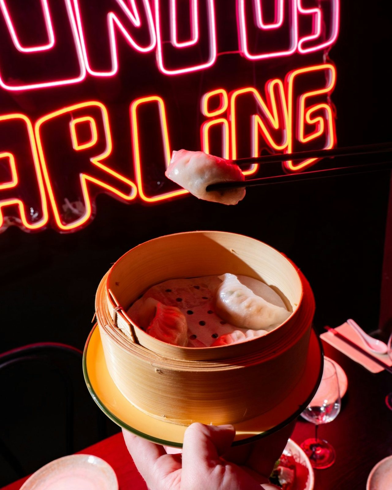 Chopsticks lifting a dumpling from a bamboo steamer in front of the Lucky You Darling neon sign